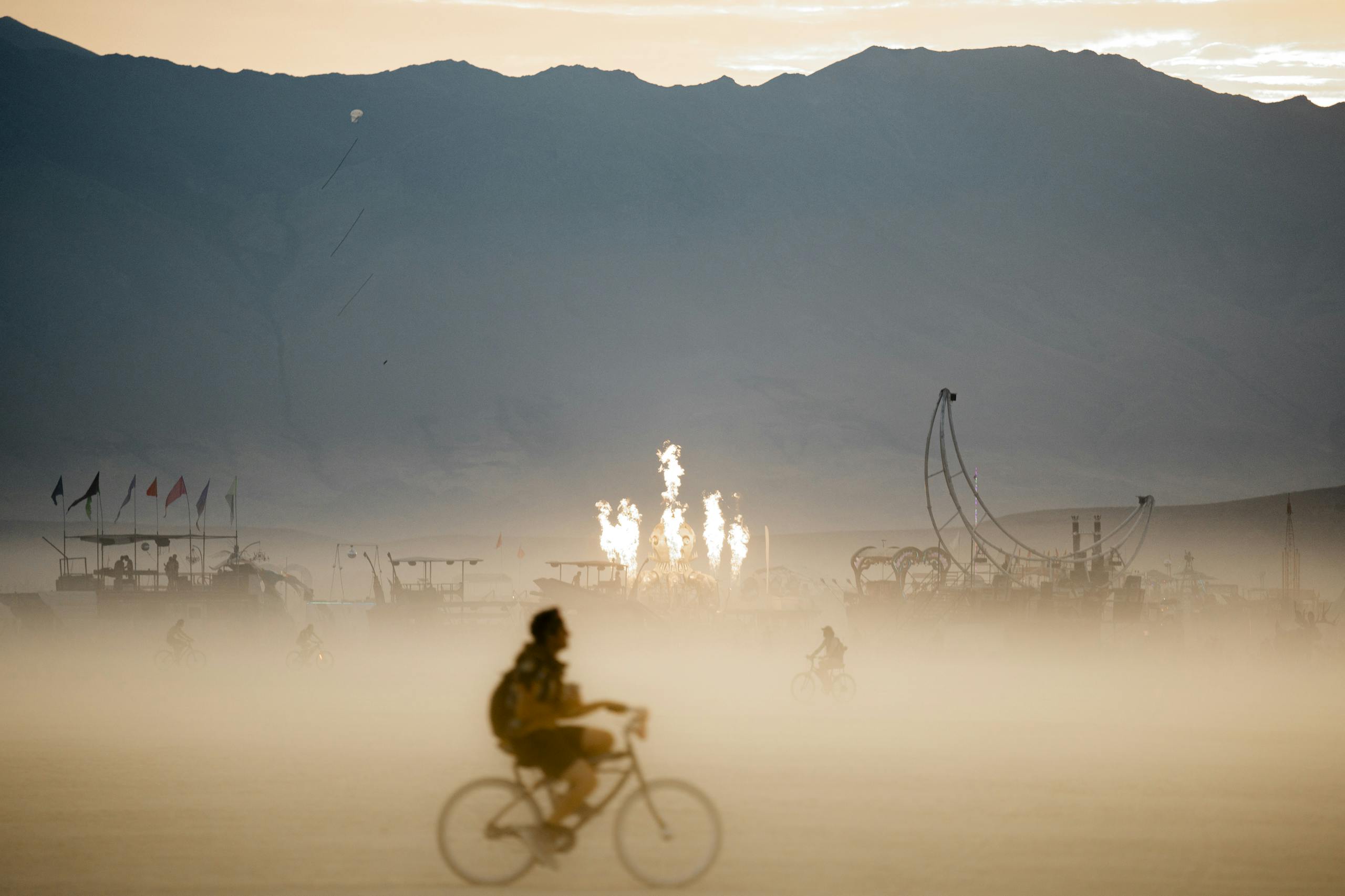 A lone cyclist rides through the desert at dusk during the Burning Man festival with art installations in the background.