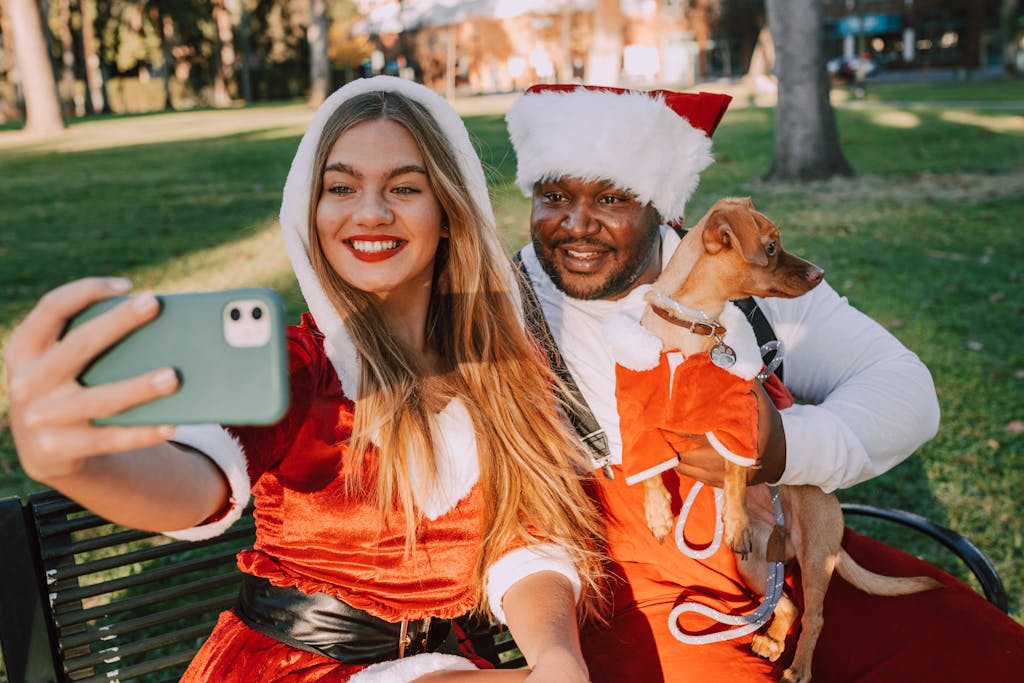 A smiling couple in Santa costumes takes a selfie with their dog on a sunny day in the park.