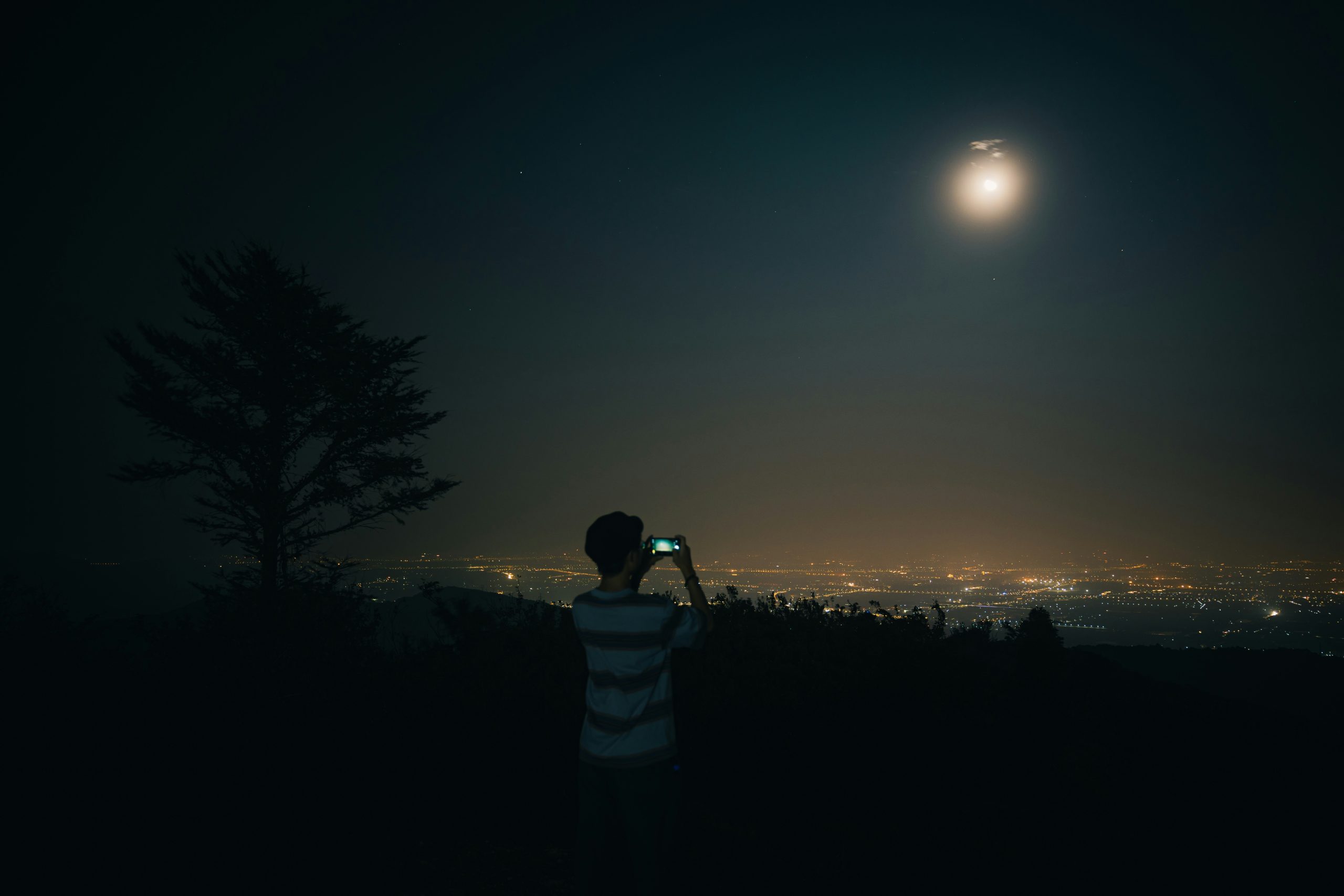 A person taking a photograph of the moon from higher ground with nighttime city lights below