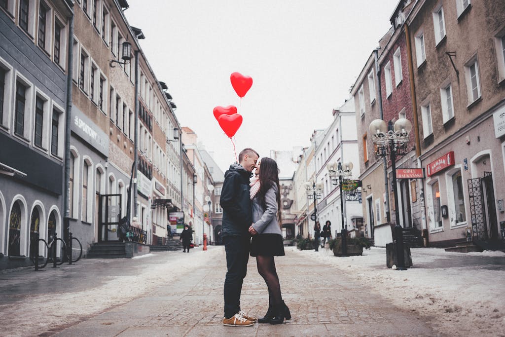A loving couple shares a kiss with heart-shaped balloons in a charming urban street setting.
