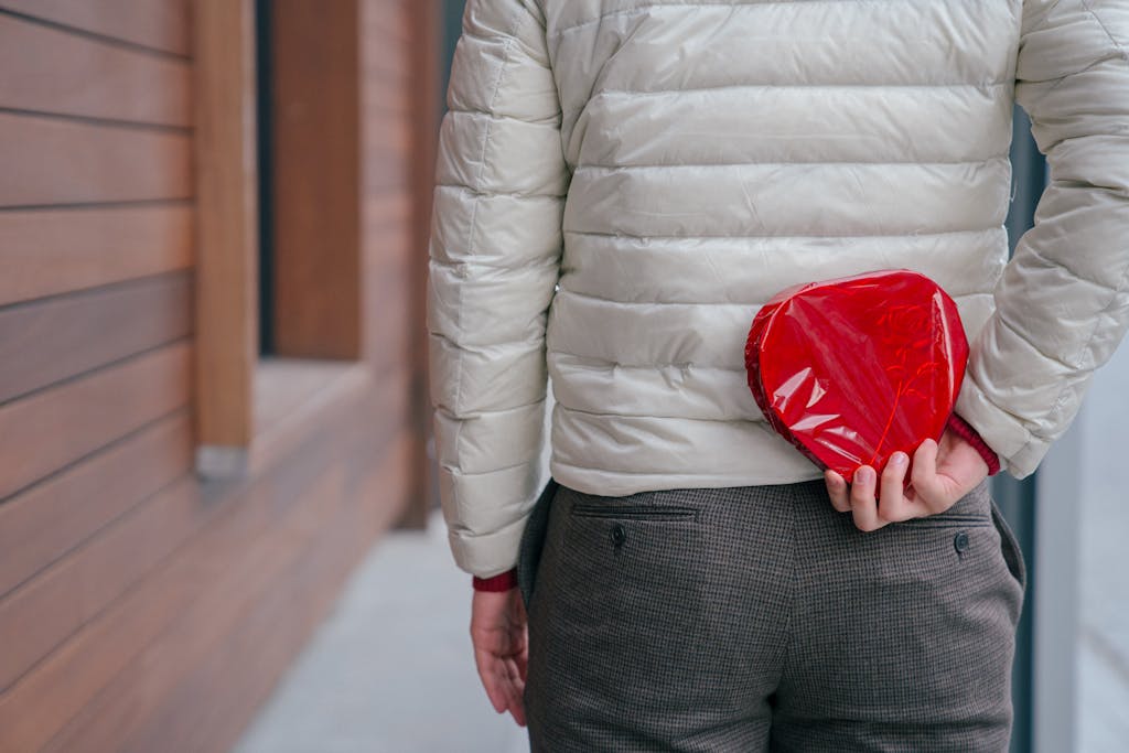 Back view of anonymous male hiding wrapped hear shaped present box while standing on street during holiday celebration in city