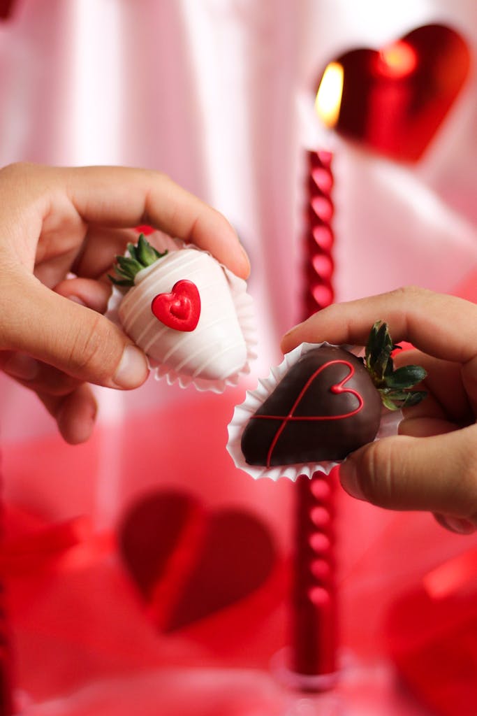 Two hands holding heart-decorated chocolate strawberries, symbolizing love.