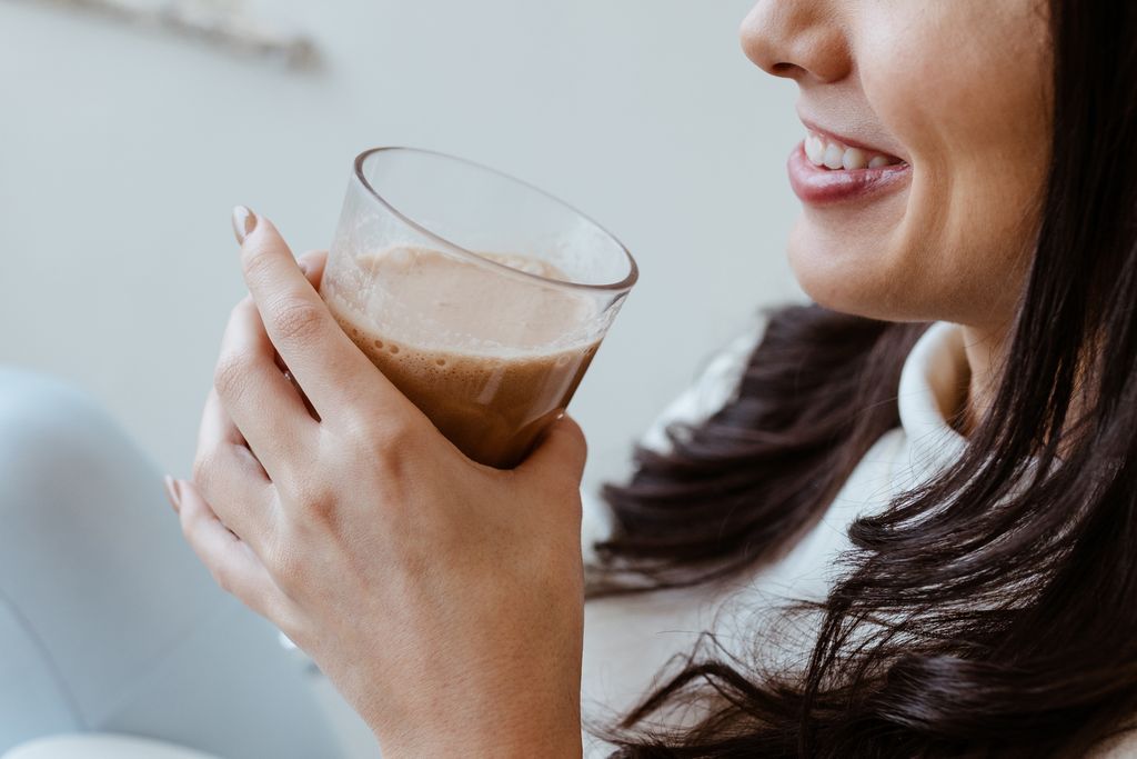 Close-up of a smiling woman enjoying a brown drink from a glass indoors. Relaxed and cozy vibe.