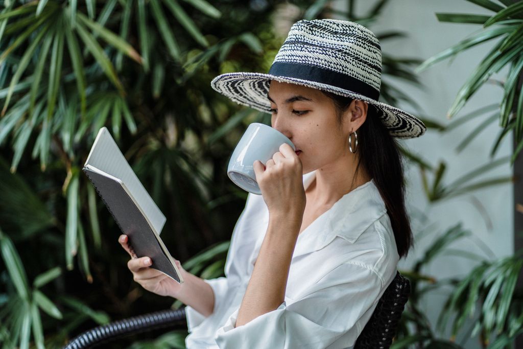 Thoughtful Asian female in stylish white shirt and straw hat reading notes and drinking cup of coffee while sitting alone near green plants